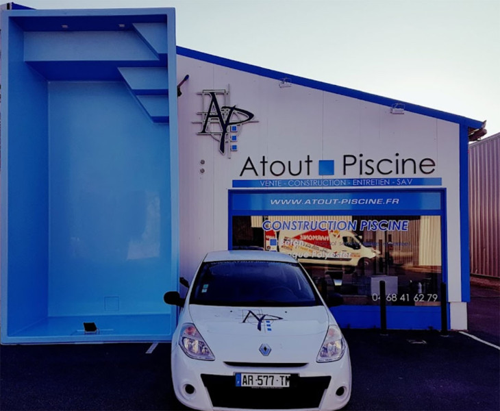 Façade du magasin Atout Piscine à Lézignan Corbière avec un grand panneau bleu affichant le modèle de piscine exposé à l'extérieur. L'enseigne du magasin indique 'Vente - Construction - Entretien - SAV' avec des informations de contact visibles, reflétant un espace professionnel dédié aux piscines.